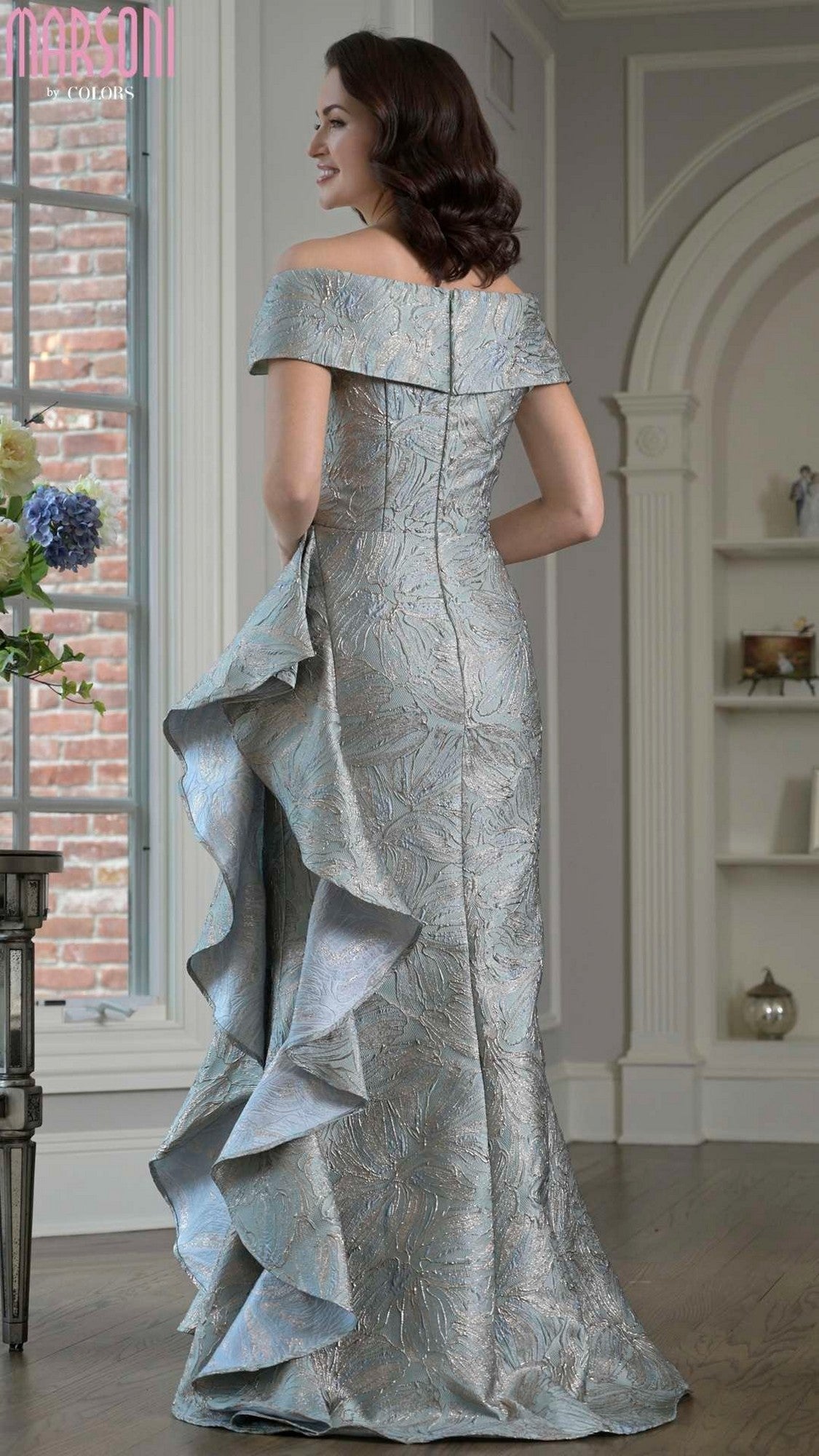 Indoors, a woman wears the Long Formal Dress: Marsoni MV1381, showcasing metallic brocade fabric, an off-the-shoulder neckline, and a cascading back ruffle detail.